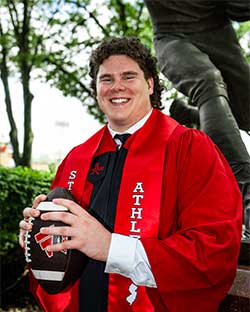 Tyler Needham in academic regalia holding a football.
