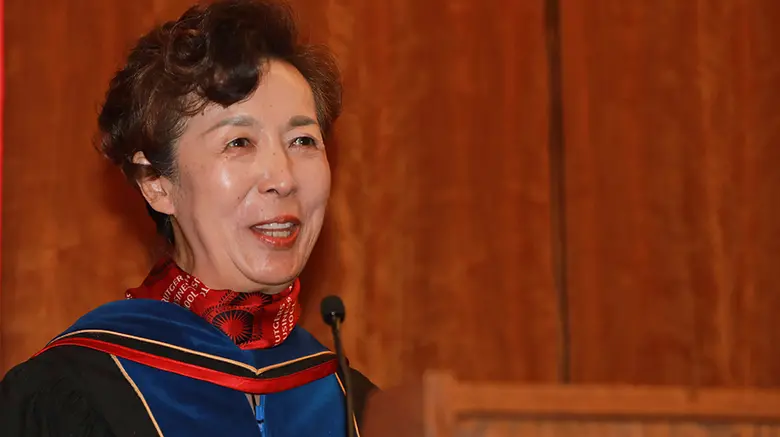 Rutgers Business School Dean Lei Lei in traditional graduation robe, addressing students at the hooding ceremony for seven Doctoral of Business Administration students.