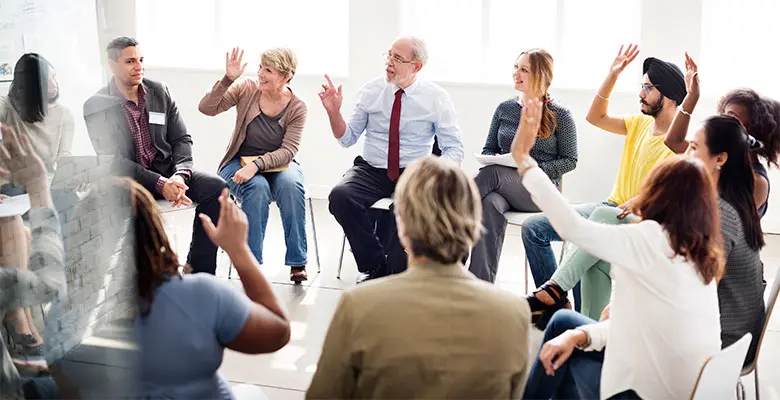 Many different types of people sit together in a circle with some raising there hands in participation
