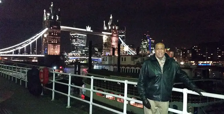 Johnny Torrance-Nesbitt, Rutgers MBA ’88 stands at River Thames at night with Tower Bridge behind him.