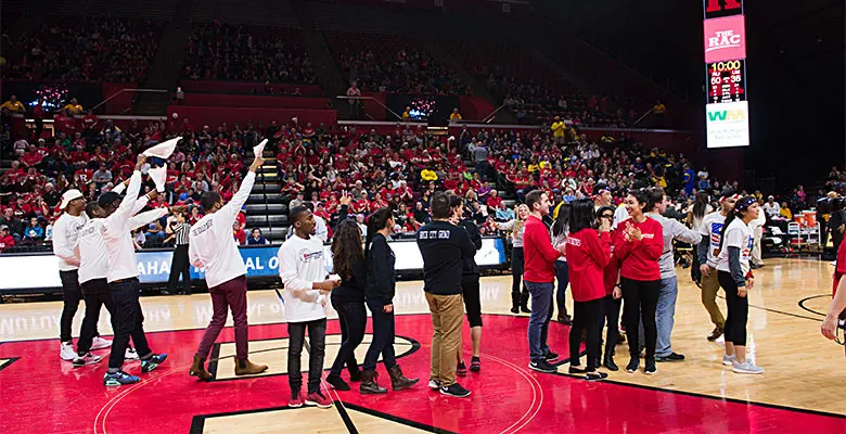 Teams lined up in the center of the basketball court with some waving their hands and towels to the crowd