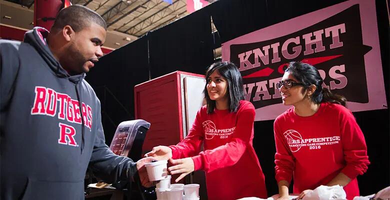 Two students pass a small white container to a fan wearing a Rutgers hoodie