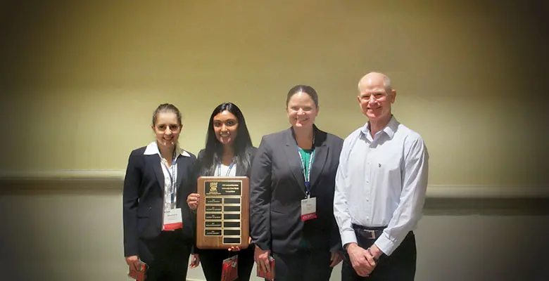 3 students appear next to Professor Goldsworthy halding a plaque of their achievements