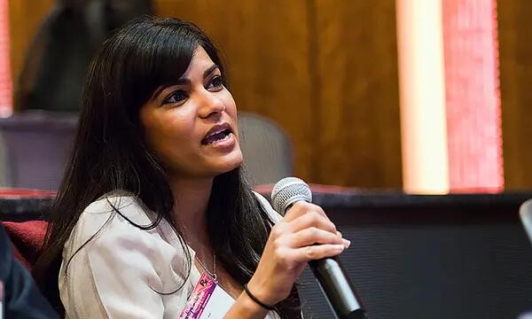 A woman with long black hair holding a microphone speaks at a past symposium