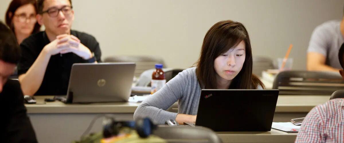 A female student's face glows in the light from her laptop as she sits in class