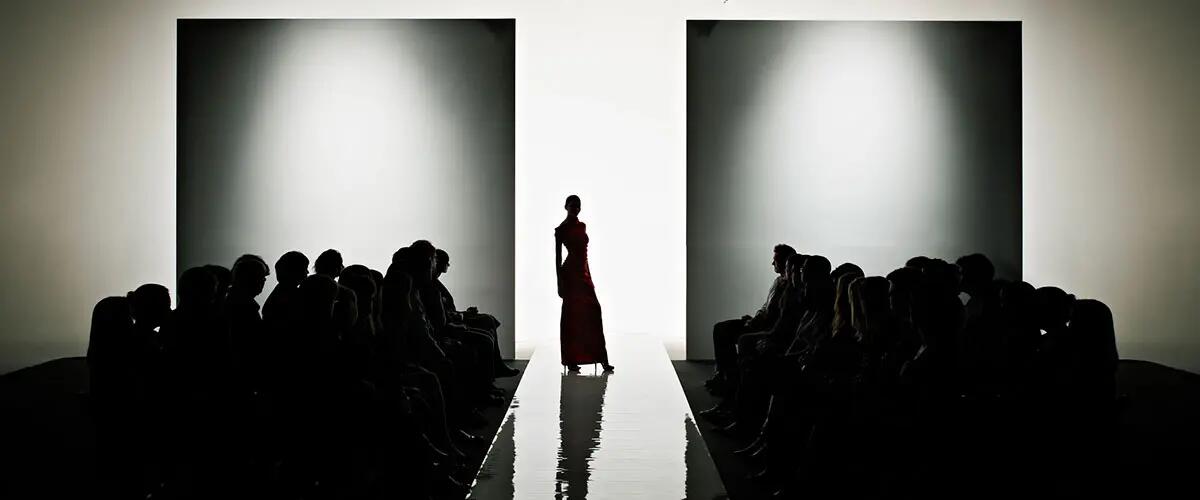 Silhouette of female model at fashion show wearing gown standing at entrance to catwalk in front of crowd